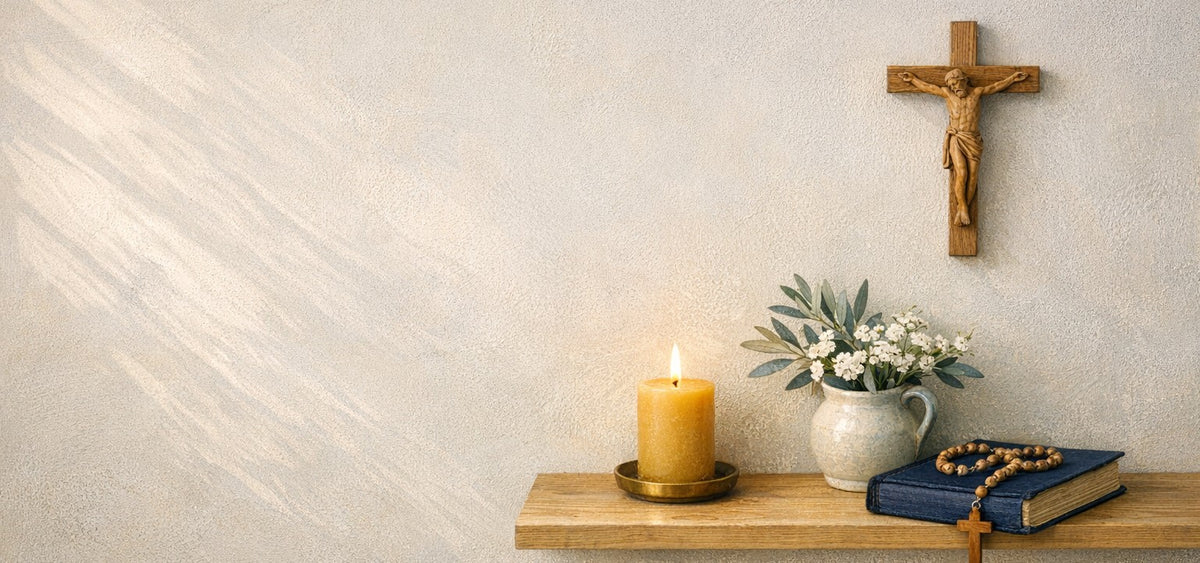 A wooden crucifix on a cream wall above a shelf with a lit beeswax candle, a vase of olive branches and white flowers, and a Bible with a wooden rosary.